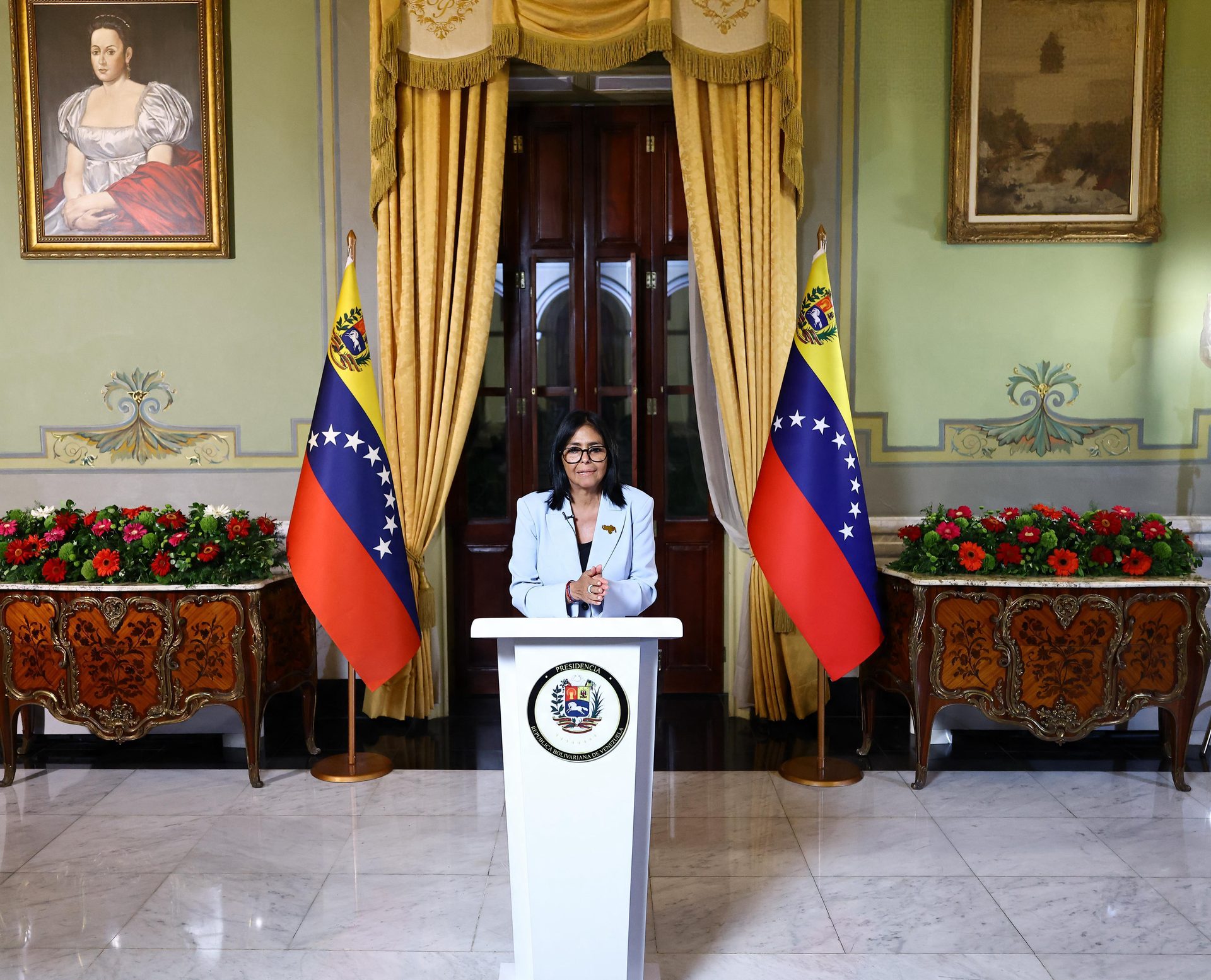 Plano general del salón del Palacio de Miraflores: la presidenta Delcy Rodríguez en el podio central entre dos banderas de Venezuela, con arreglos florales y pinturas al fondo.