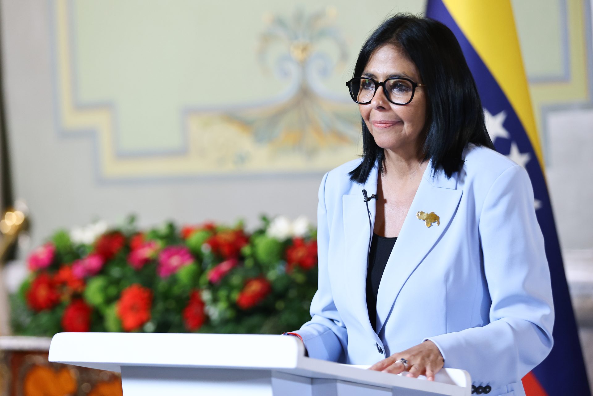 La presidenta Delcy Rodríguez ante el podio, en plano de tres cuartos, con un arreglo floral y la bandera de Venezuela al fondo.